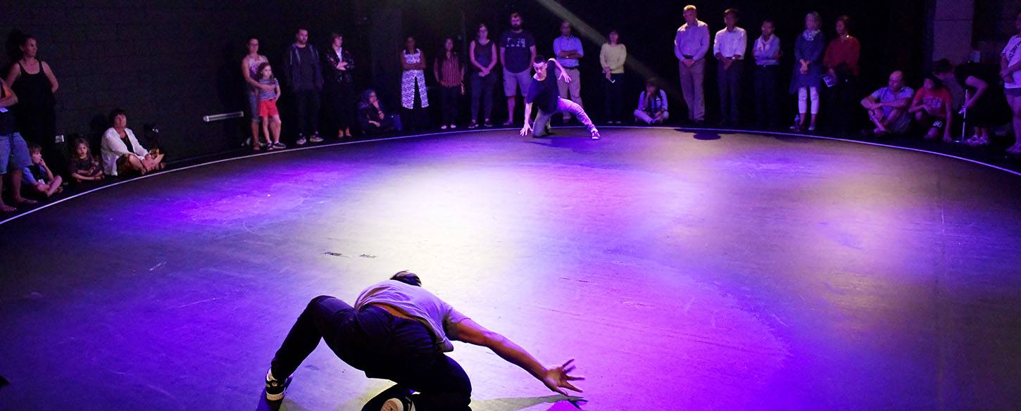 Two men are at opposite ends of a stage and crouching on the floor about to dance battle with an audience surrounding them