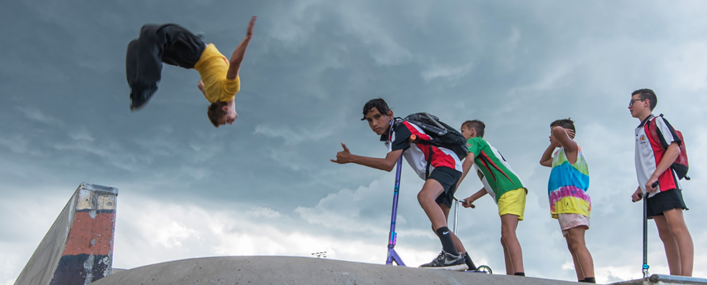 group of young boys stand on a ledge in a skate park in a row whilst a man does a backflip