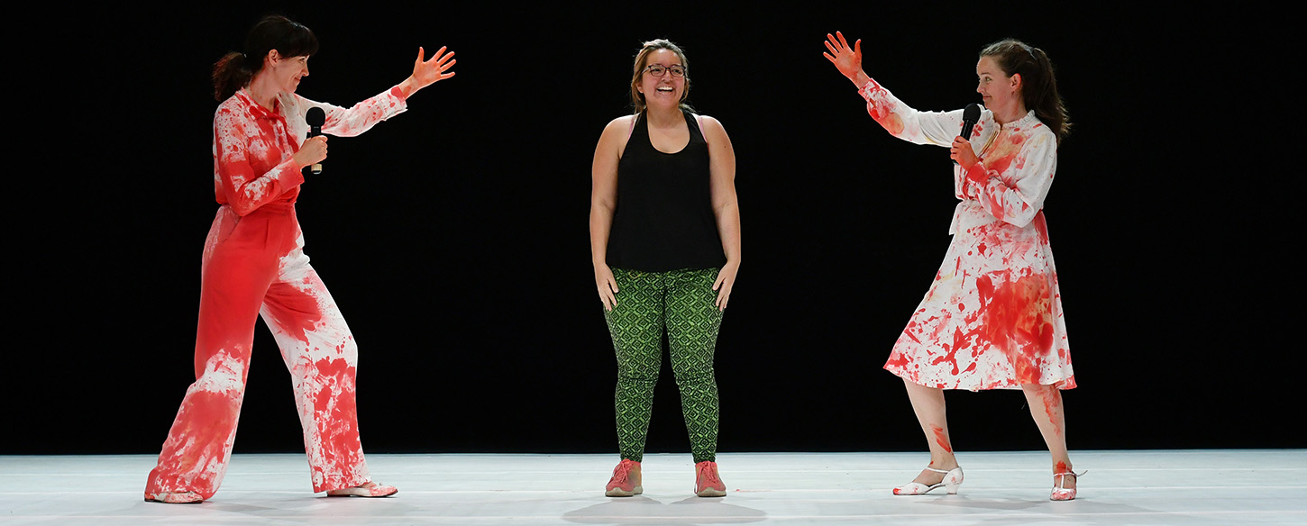 Two women performers in white suits with blood stand either side an audience member in the centre