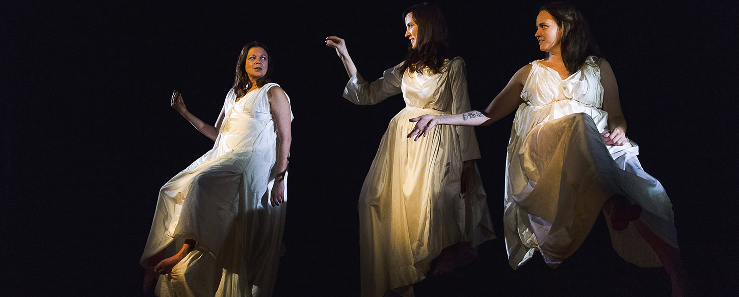 Three women wearing white gowns sit on high stools against a black backdrop