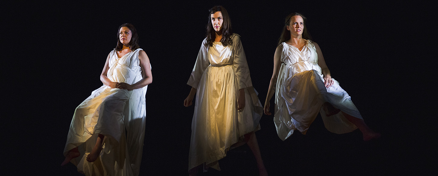 Three women wearing white gowns sit on high stools against a black backdrop