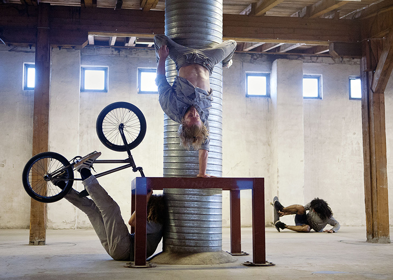 three performers are in an industrial space - one holds a bike, the other is doing a handstand and another is holding a skateboard