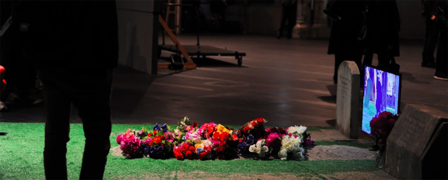 a tv and a fake grass with flowers are on a floor and audience looks on